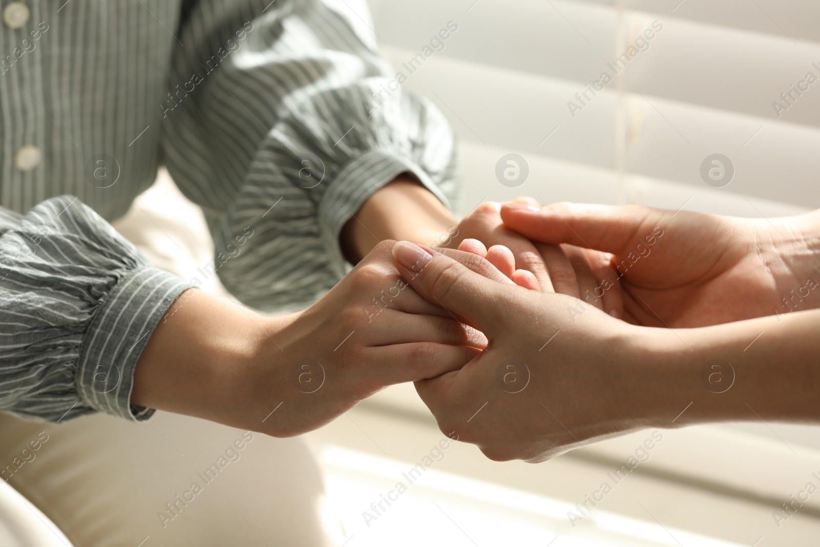 Photo of Religious women holding hands and praying together near window indoors, closeup