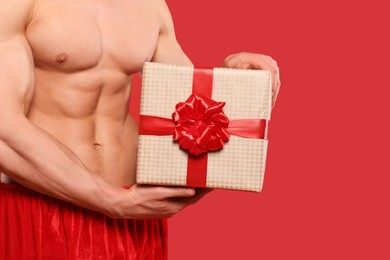 Young man with muscular body holding Christmas gift box on red background, closeup. Space for text