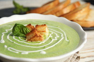 Photo of Bowl of delicious broccoli cream soup with croutons and basil on table, closeup