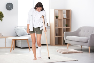 Young woman with crutch and broken leg in cast at home