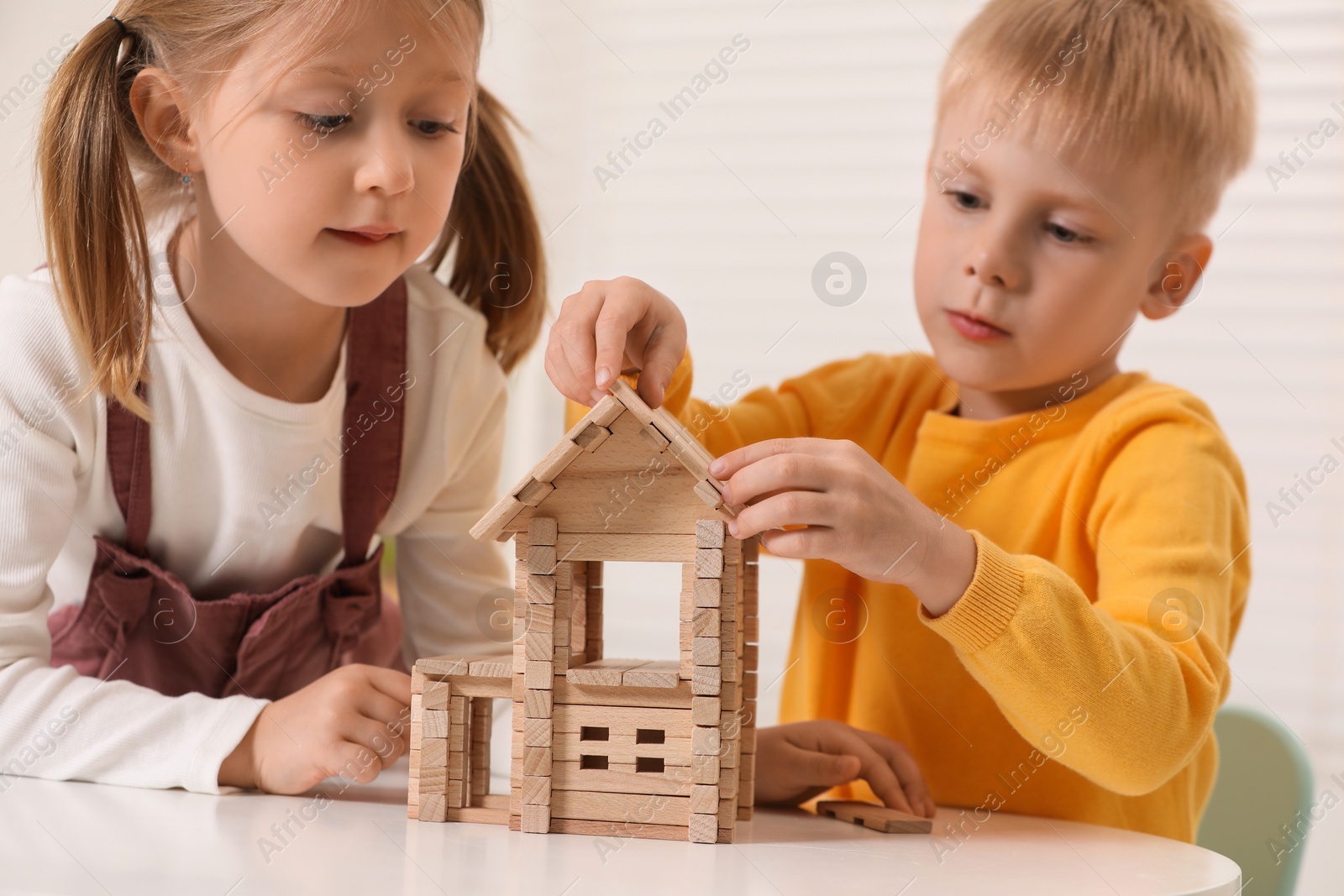 Photo of Little boy and girl playing with wooden house at white table indoors. Children's toys