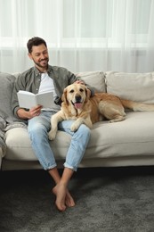 Photo of Man reading book on sofa near his cute Labrador Retriever at home