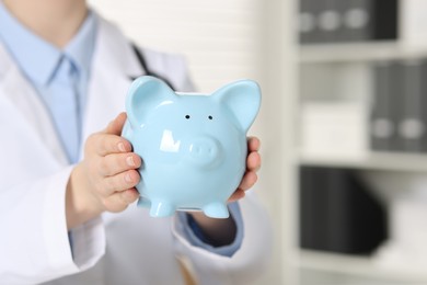 Photo of Doctor with light blue piggy bank indoors, closeup. Space for text