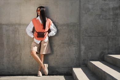 Photo of Beautiful young woman with stylish waist bag near grey wall outdoors, space for text