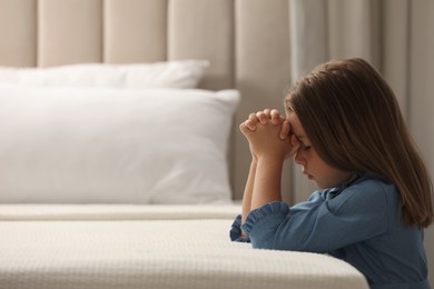 Cute little girl with hands clasped together saying bedtime prayer in bedroom. Space for text