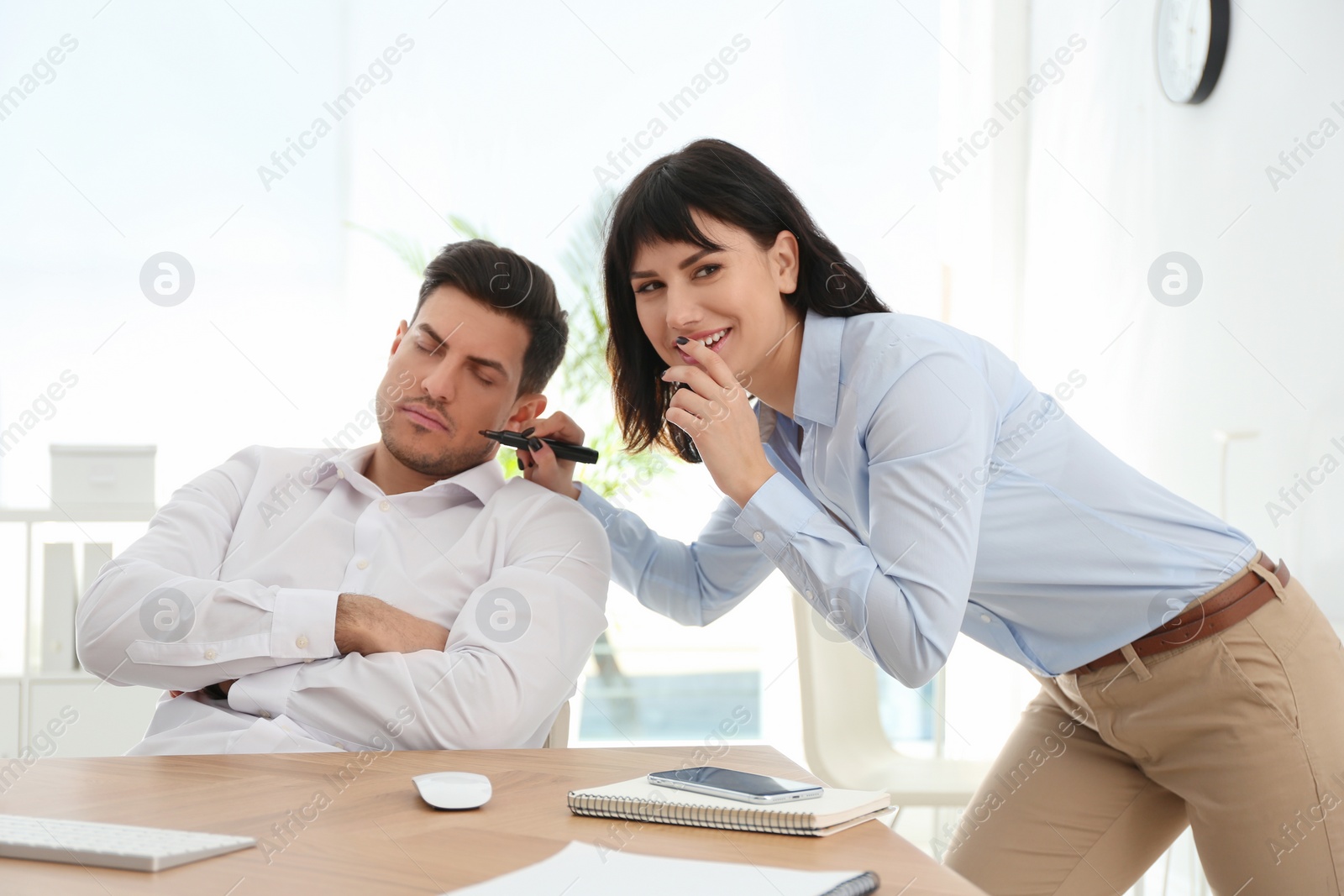 Photo of Young woman drawing on colleague's face while he sleeping at workplace. April fool's day