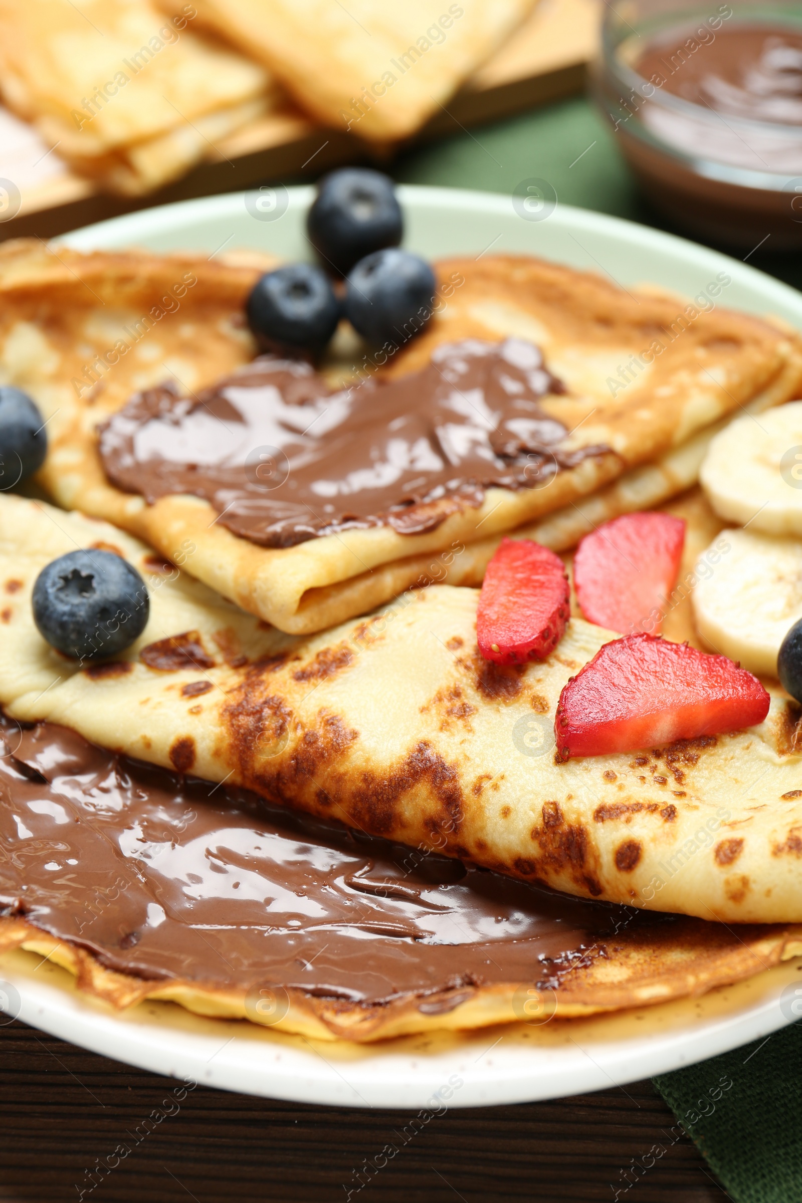 Photo of Tasty crepes with chocolate paste, banana and berries on table, closeup