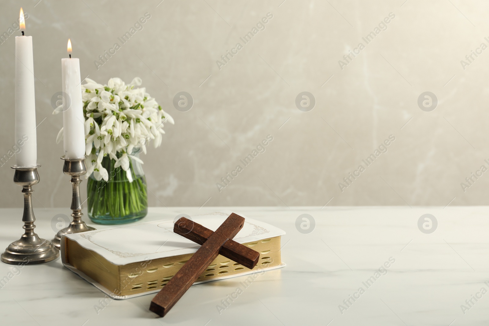 Photo of Burning church candles, wooden cross, Bible and flowers on white marble table. Space for text