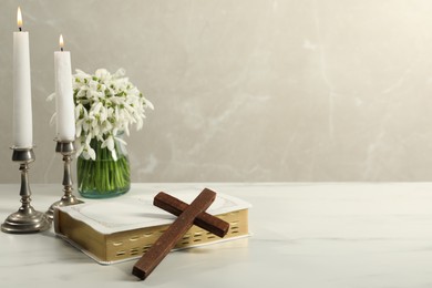Burning church candles, wooden cross, Bible and flowers on white marble table. Space for text