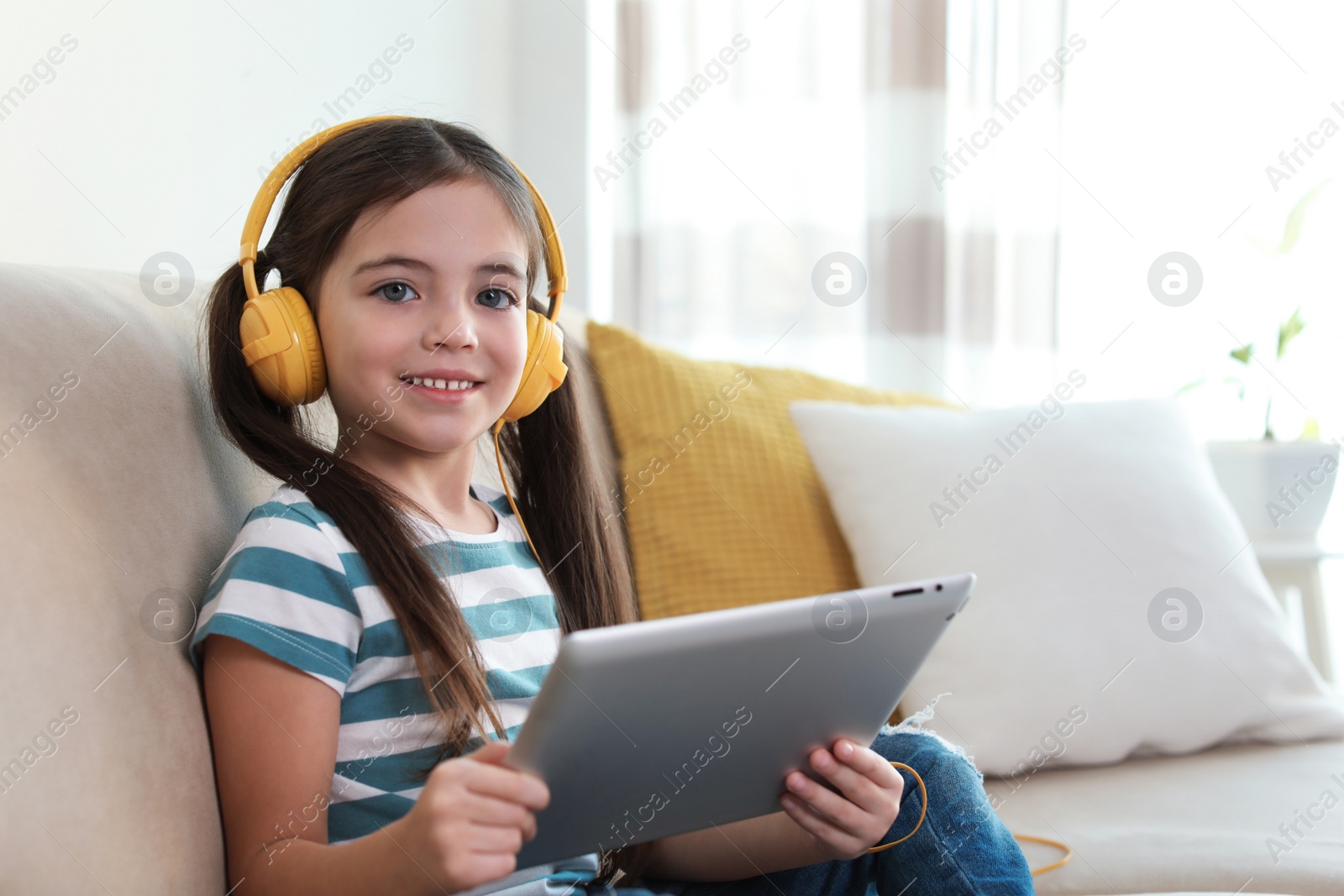 Photo of Cute little girl with headphones and tablet listening to audiobook at home