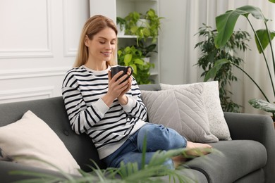Photo of Woman with cup of drink sitting on sofa surrounded by beautiful potted houseplants at home