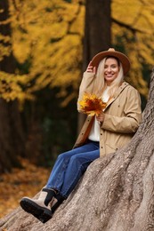 Photo of Happy woman with dry autumn leaves sitting on tree trunk outdoors