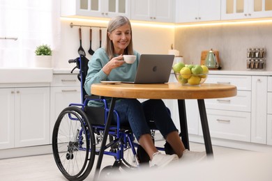 Photo of Woman in wheelchair with cup of drink using laptop at table in home office