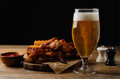 Photo of Glass of beer, delicious baked chicken wings, grilled corn and sauce on wooden table against black background