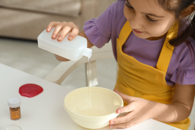 Cute little girl pouring glue into bowl at table in room. DIY slime toy