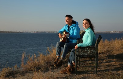 Couple with guitar resting in camping chairs near river on sunny day