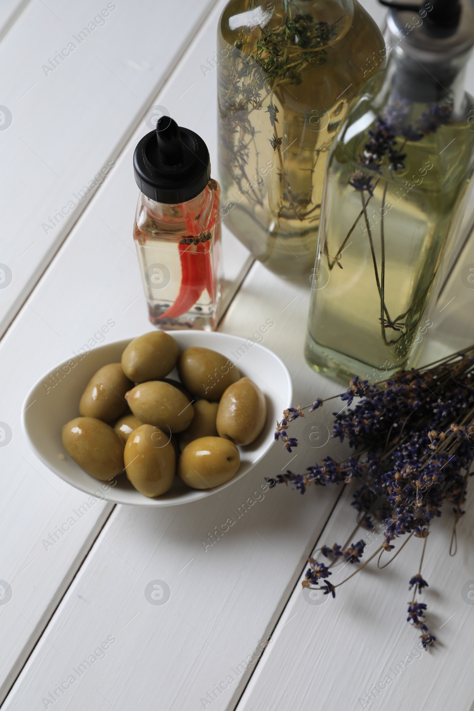 Photo of Bottles of different cooking oils, lavender flowers and olives on white wooden table