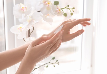 Photo of Young woman applying cream onto hands against window