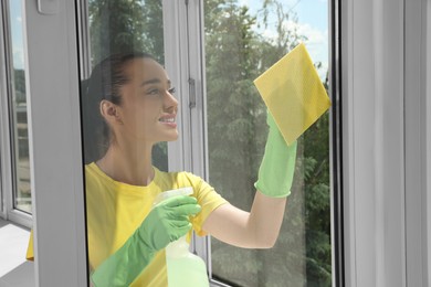 Happy young woman cleaning window glass with sponge cloth and spray indoors