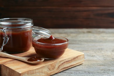 Tasty barbeque sauce in bowl, jar and spoon on wooden table, closeup. Space for text