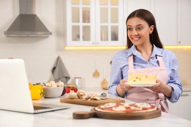 Happy woman making pizza while watching online cooking course via laptop in kitchen