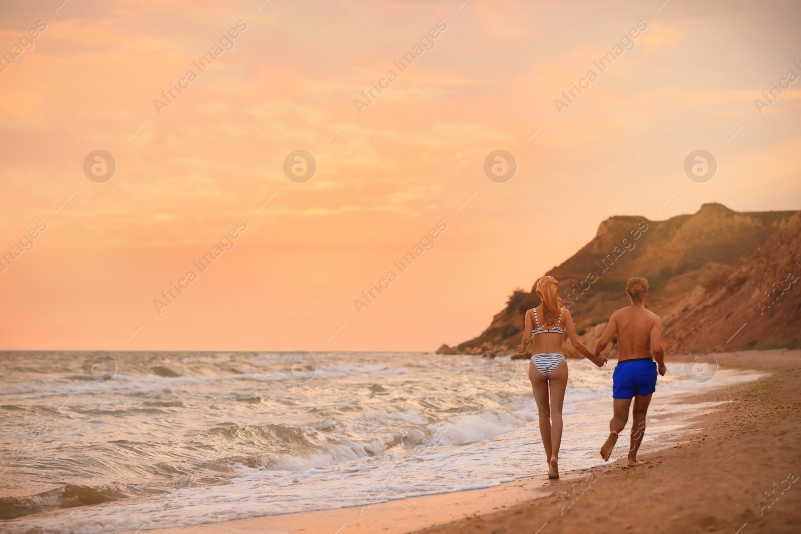 Photo of Young woman in bikini and her boyfriend walking on beach at sunset. Lovely couple