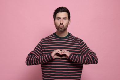 Handsome man making heart with hands and blowing kiss on pink background