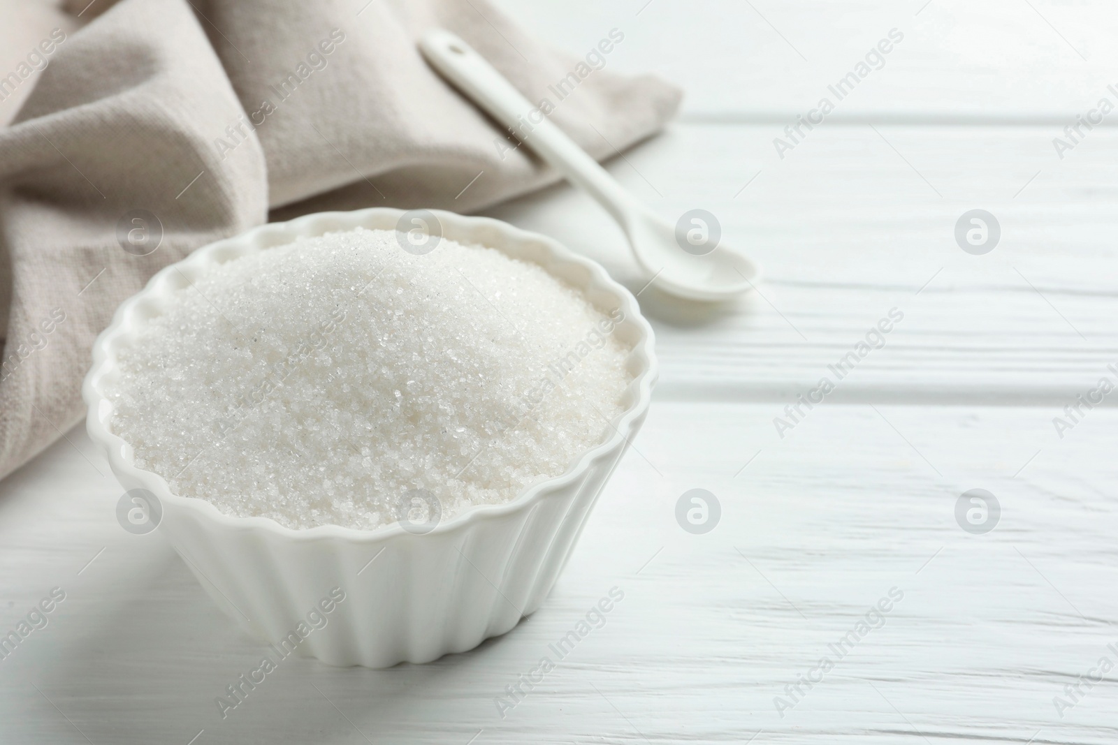 Photo of Granulated sugar in bowl on white wooden table, closeup. Space for text