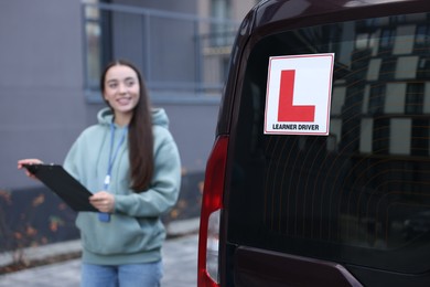 Photo of Instructor with clipboard near car outdoors, selective focus on L-plate. Driving school