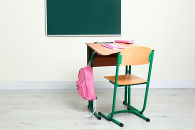 Photo of Wooden school desk with stationery and backpack near chalkboard in classroom