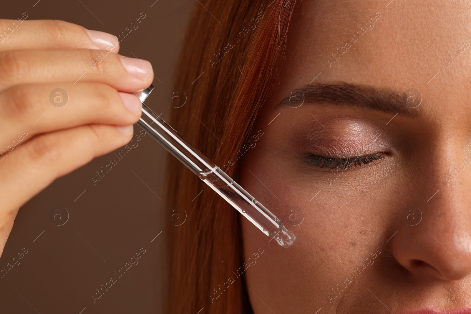 Photo of Beautiful young woman applying cosmetic serum onto her face on brown background, closeup