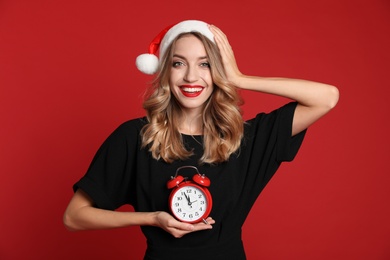 Photo of Happy young woman wearing Santa hat with alarm clock on red background. Christmas time