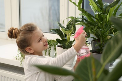 Photo of Cute little girl spraying beautiful green plant on windowsill at home. House decor