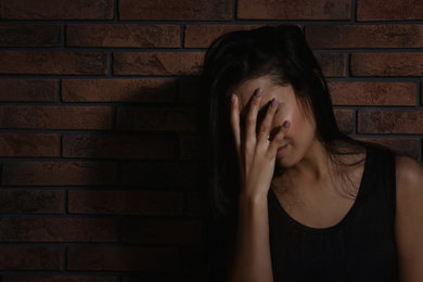 Photo of Crying young woman near brick wall, space for text. Domestic violence concept