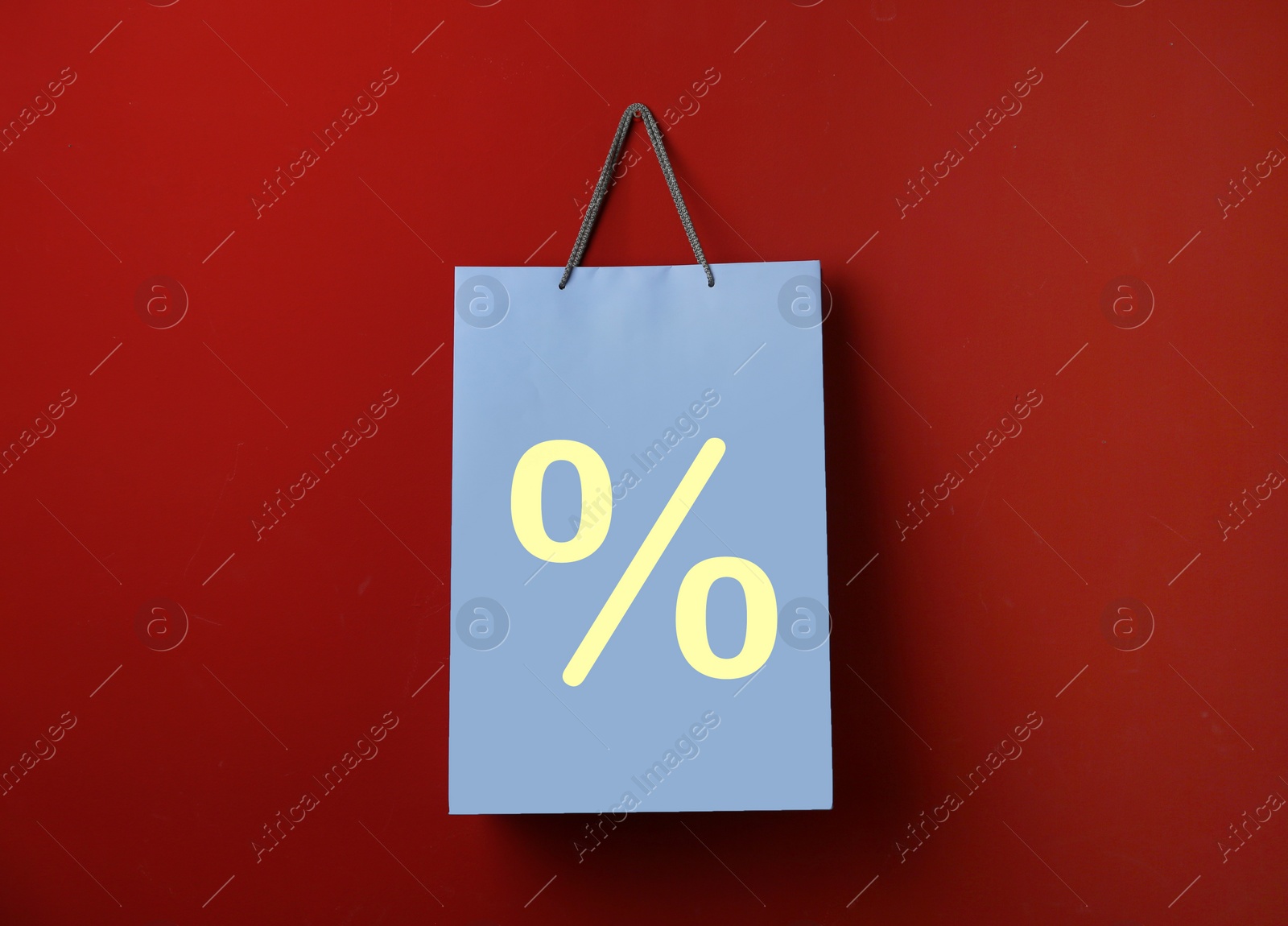 Image of Light blue paper shopping bag with percent sign hanging on red wall. Discount concept
