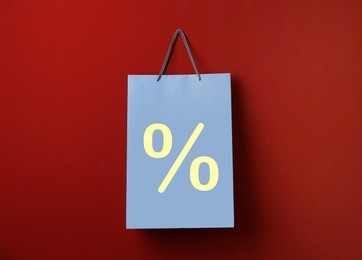 Light blue paper shopping bag with percent sign hanging on red wall. Discount concept