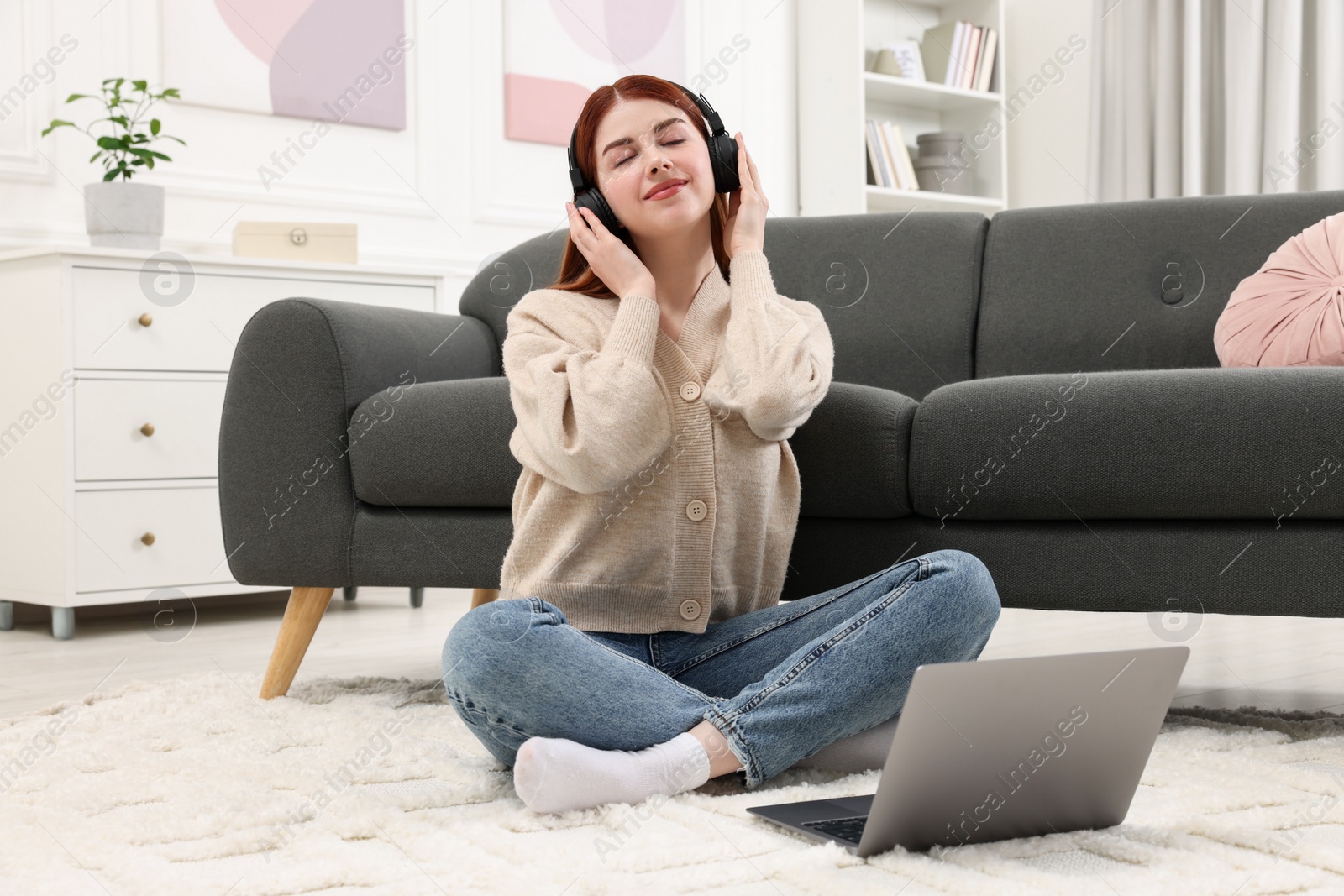 Photo of Beautiful woman with headphones listening to music near laptop on rug in living room