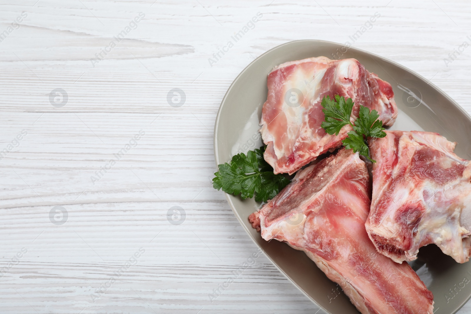 Photo of Plate with raw chopped meaty bones and parsley on white wooden table, top view. Space for text