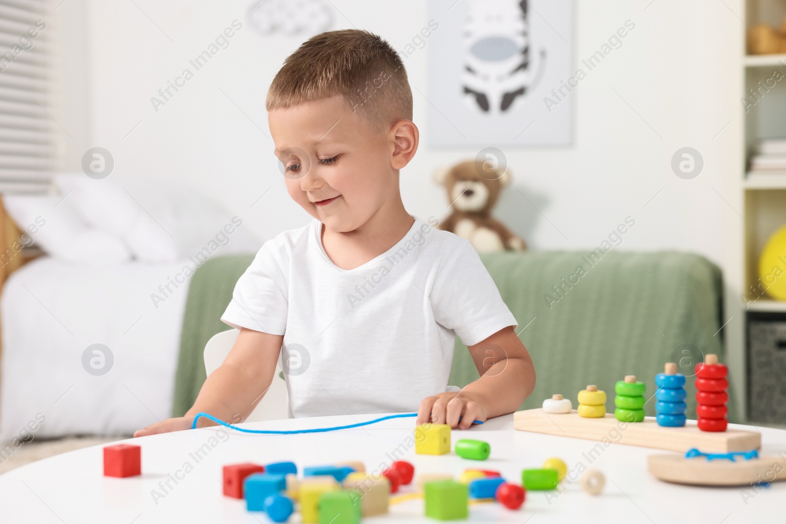 Photo of Motor skills development. Little boy playing with wooden pieces and string for threading activity at white table indoors