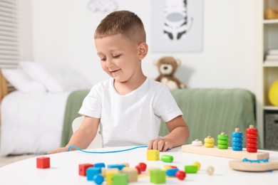 Photo of Motor skills development. Little boy playing with wooden pieces and string for threading activity at white table indoors