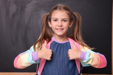 Photo of Smiling schoolgirl showing thumbs up near blackboard