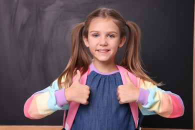 Smiling schoolgirl showing thumbs up near blackboard