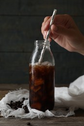 Woman stirring delicious iced coffee with straw in glass bottle at wooden table, closeup