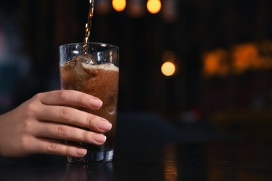 Woman holding glass with ice cubes while pouring cola at table indoors, closeup. Space for text