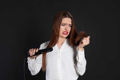 Stressed young woman with flattening iron on black background. Hair damage