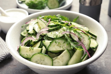 Photo of Delicious cucumber salad with onion and arugula in bowl on grey table, closeup