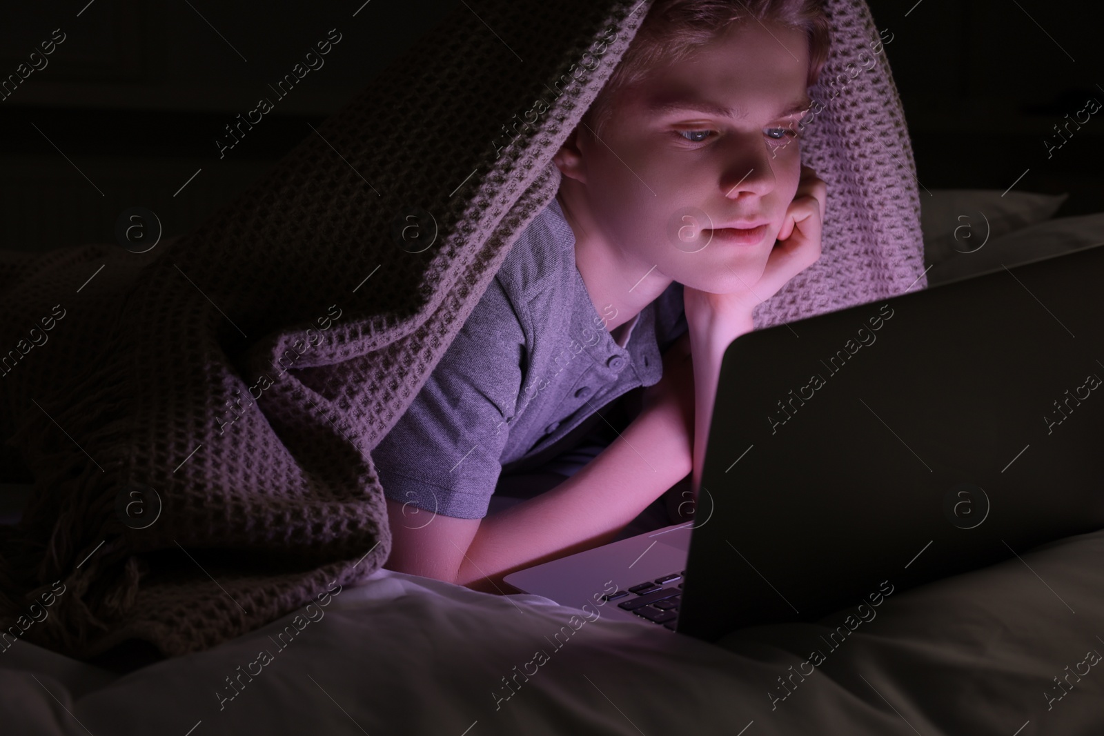 Photo of Teenage boy using laptop under blanket on bed at night. Internet addiction