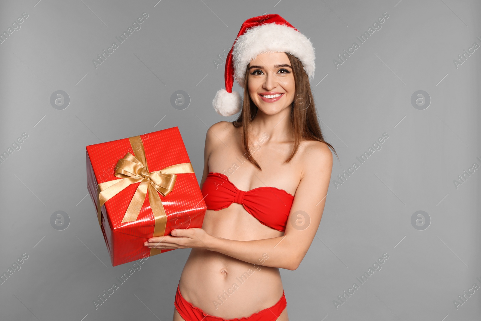 Photo of Happy young woman in red bikini and Santa hat with gift box on light grey background. Christmas celebration