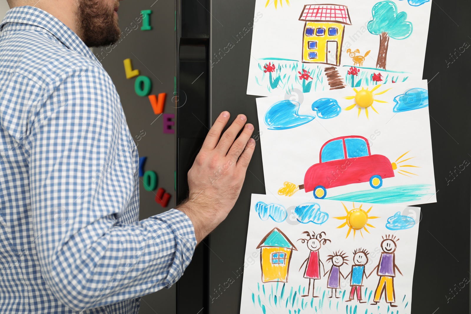 Photo of Man opening refrigerator door with child's drawings and magnets, closeup