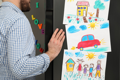 Photo of Man opening refrigerator door with child's drawings and magnets, closeup